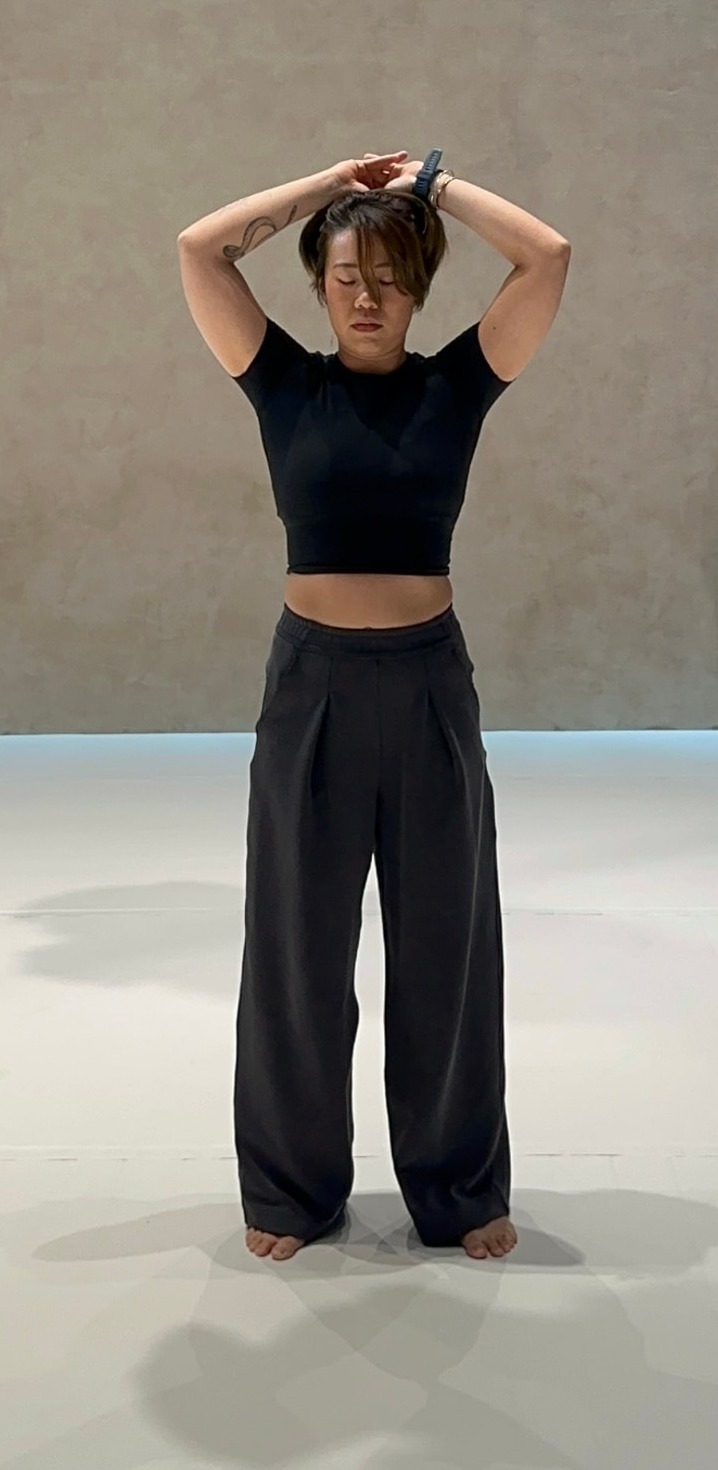 Sharone standing barefoot in a studio, arms folded overhead, eyes closed